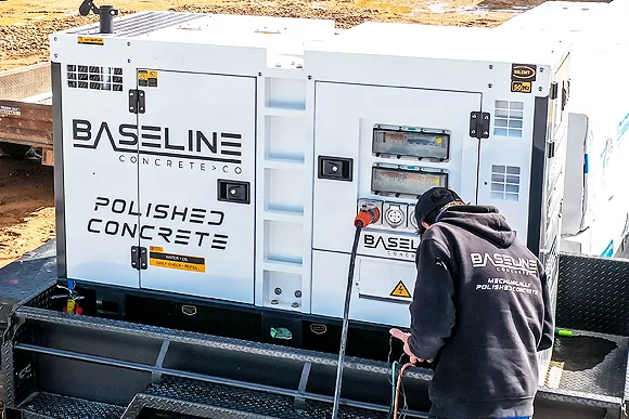 A worker connects equipment to a Baseline Concrete Co. generator, marked with "Polished Concrete," at a construction site.