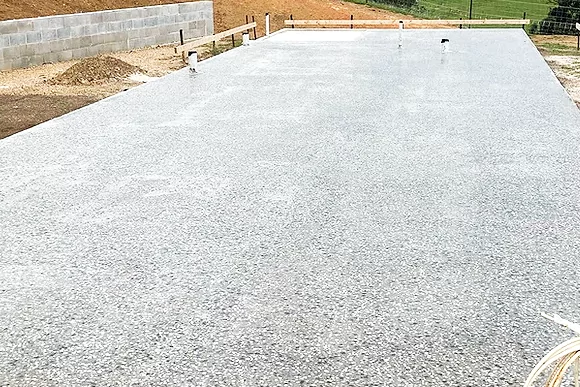A smooth, newly poured concrete slab sits on a construction site, edged by a gray block wall and a landscaped area