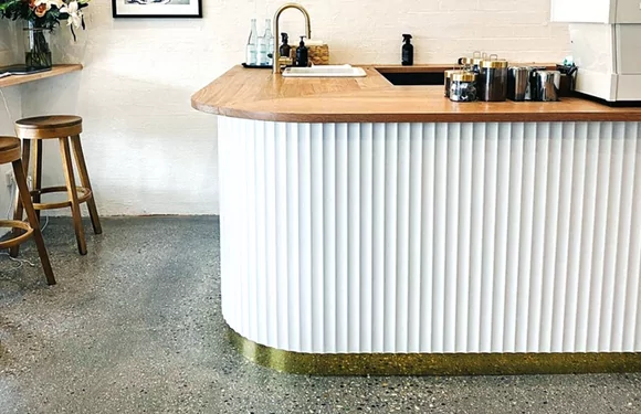 A modern bar area featuring a curved white and gold countertop with wooden top, flanked by wooden stools and minimalist decor.
