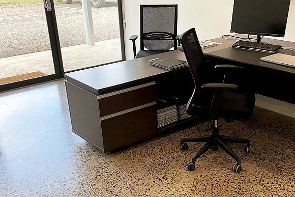 A modern office space featuring a dark wooden desk, ergonomic chair, and computer setup, with a view of an outdoor area.