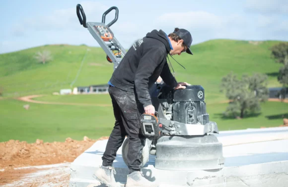 A worker operates a concrete finisher on a construction site, with green hills and a building in the background