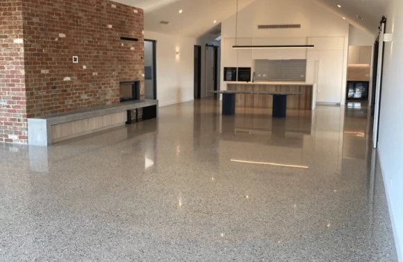 Spacious modern living area with high ceilings, exposed brick wall, kitchen in the background, and polished terrazzo flooring