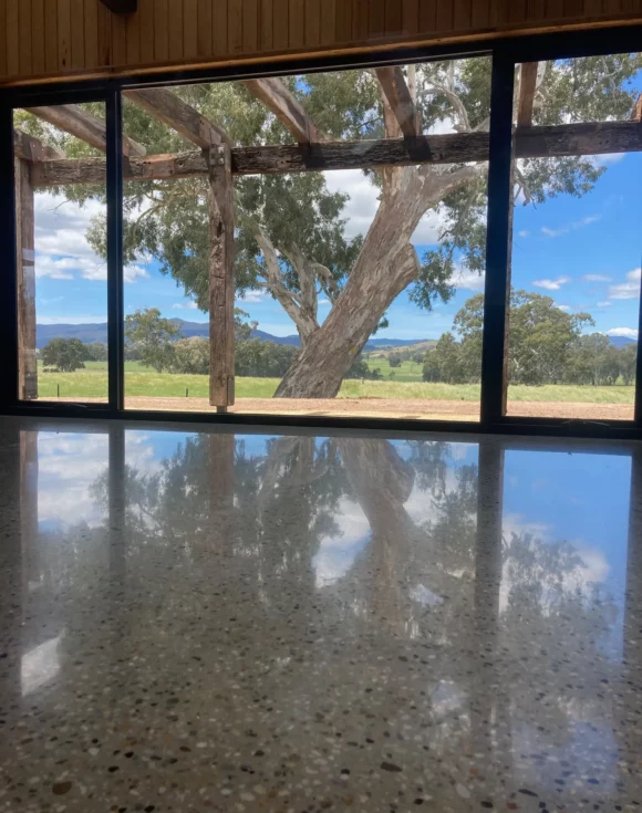 View through large windows of a rural landscape with trees and distant hills. A tree is reflected on the polished concrete floor inside.