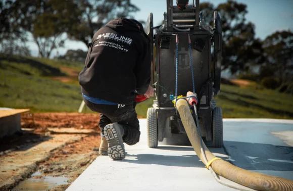 A worker in a hoodie kneels next to a concrete machine, guiding a hose on a concrete slab amidst a green landscape