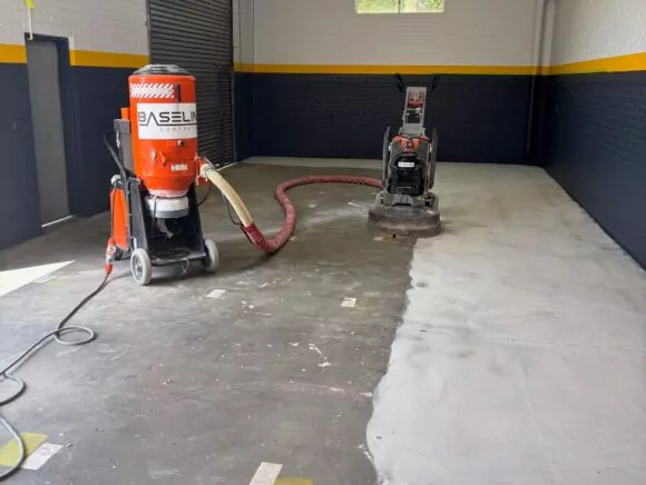 A construction site with a concrete floor being sanded by a machine, alongside a vacuum for dust collection. Yellow stripe on walls.
